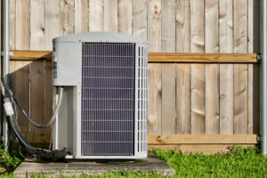 Central air conditioning unit in a residential backyard in front of a defocused wooden fence and lawn area